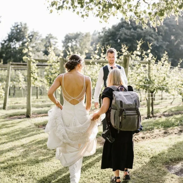Bride in a wedding dress walking through a vineyard with a person carrying a backpack.