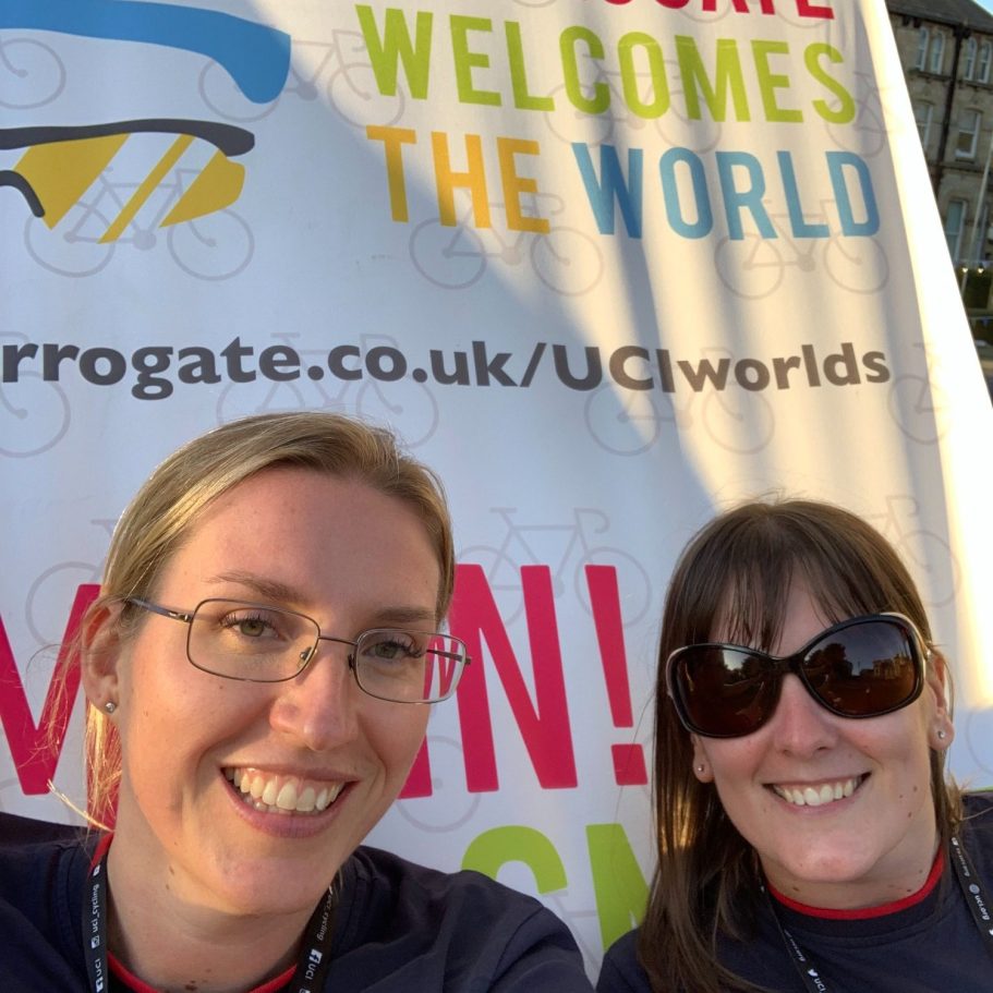 Two women smiling in front of a banner that reads "Harrogate Welcomes the World".