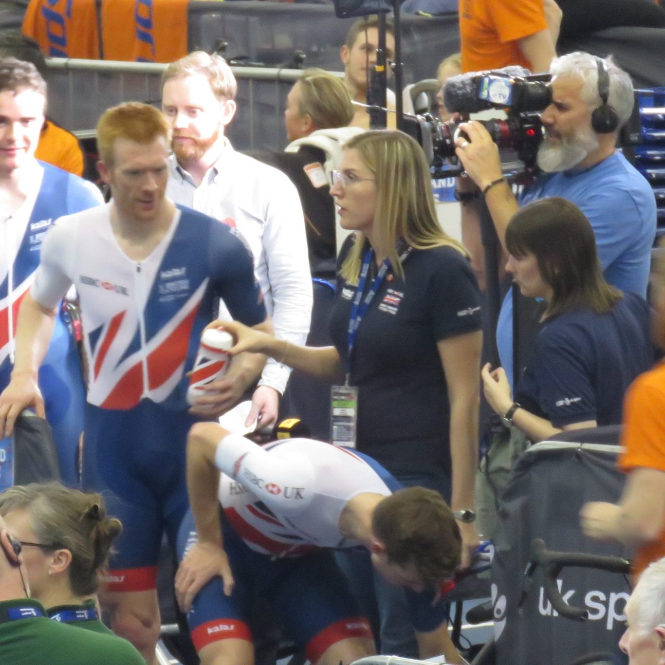 Athletes in cycling uniforms converse and prepare before a race, with a media crew nearby.